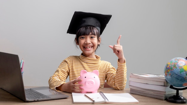 ピンクの貯金箱で卒業帽をかぶったアジアの少女将来のお金の投資を節約