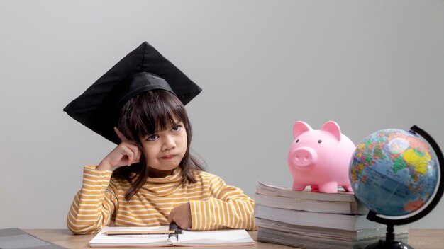 Asian little girl wearing a graduation cap with a pink piggy bank Saving money investment the future