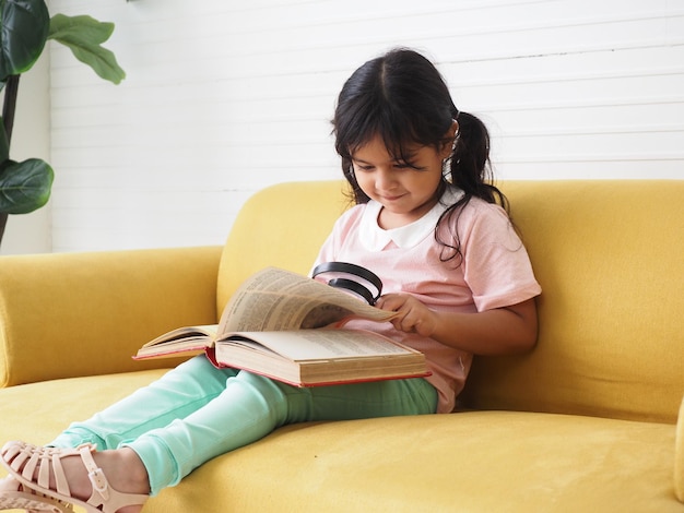 Photo an asian little girl with glasses is sitting on a yellow sofa at home and reading a book with a smart funny expression on her face learning conceptxa