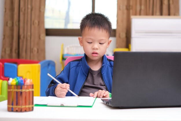 아시아 작은 아이가 집에서 온라인 수업 중 숙제를 공부하고 노트북 컴퓨터를 사용하여 그리기