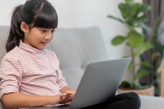 아시아계 어린 학교 소녀는 집에서 혼자 소파에 앉아 노트북 컴퓨터를 사용합니다.
