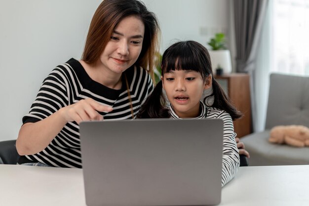 Asian little young girl kid learning online class at home with mother Preschool child use laptop computer do homework homeschool from school teacher by digital remote internet with support from mom