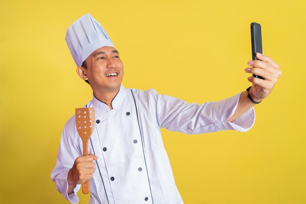 Asian male chef wearing chef jacket selfie while holding spatula on isolated background