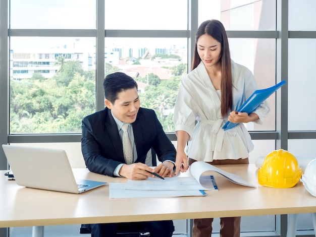 Asian male chief architect or engineer in suit and young female secretary discuss on blueprint with laptop computer and white and yellow hard hats on desk on huge glass window in office.