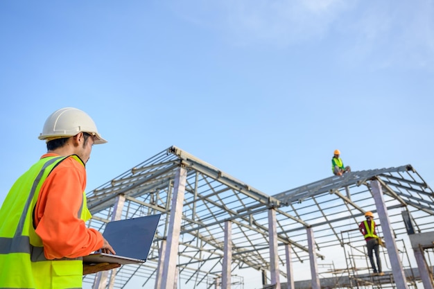 Asian male construction engineer holding a laptop standing at a house construction site Controlling and ordering Workers building houses and steel roof trusses