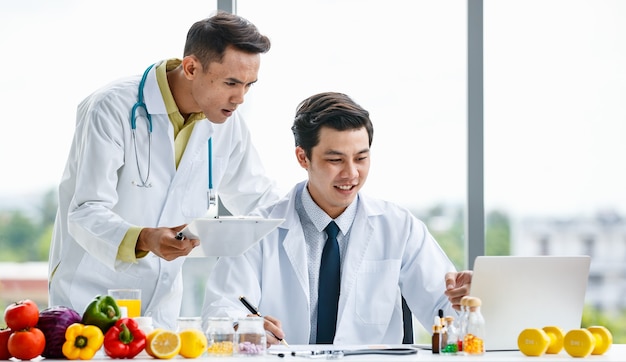 제복을 입은 아시아 남성 의사들은 함께 병원에서 일하는 동안 과일과 비타민 근처에 있는 클립보드와 노트북에서 데이터를 분석하고 웃고 있습니다.