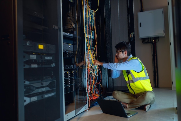 コンピューターのサーバールームで検査しながら、ラップトップを持ったアジア人の男性エンジニアがスマートフォンで話している。データベース サーバーの管理とメンテナンスの概念