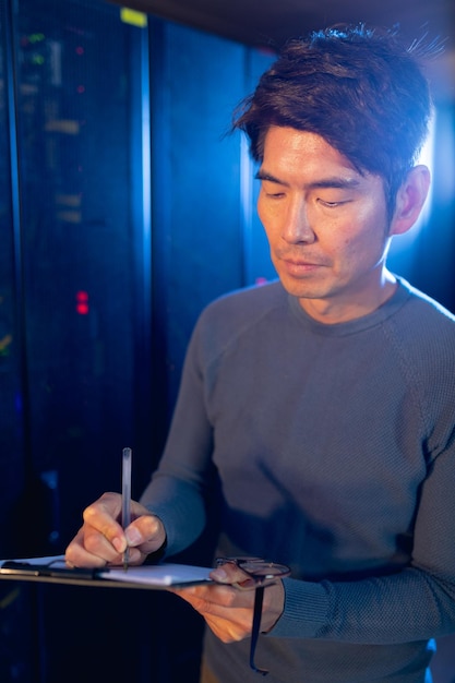 Asian male engineer writing on clipboard in computer server room. database server management and maintenance concept