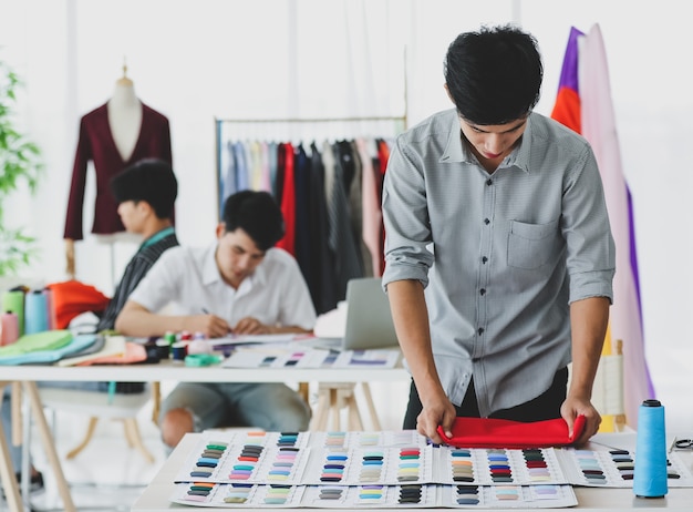 Asian male fashion designer comparing fabric with colorful samples while working near colleagues in atelier