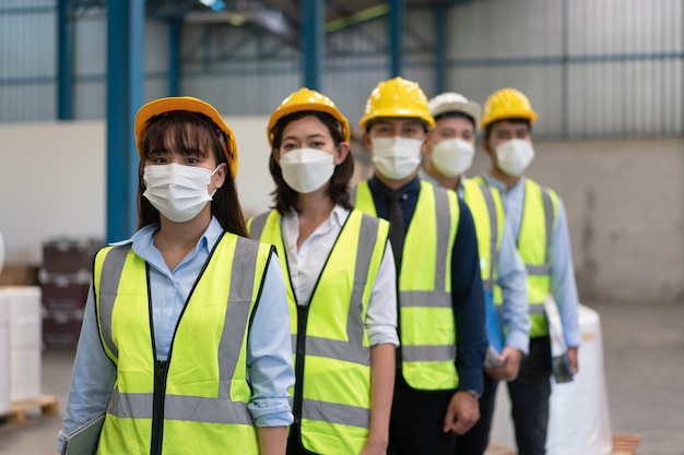 Il team di ingegneri asiatici maschi e femmine indossa una maschera con supporto di sicurezza del casco in linea nella fabbrica del magazzino