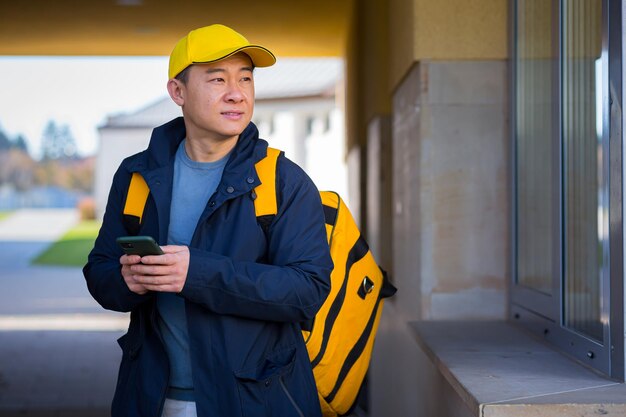 Asian male food courier using mobile using mobile phone app on\
smartphone while standing city street with insulated with thermal\
backpack delivering food looking customer address purchase online\
map