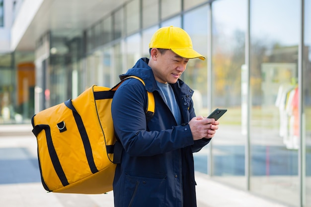 Asian male food courier using mobile using mobile phone app on\
smartphone while standing city street with insulated with thermal\
backpack delivering food looking customer address purchase online\
map