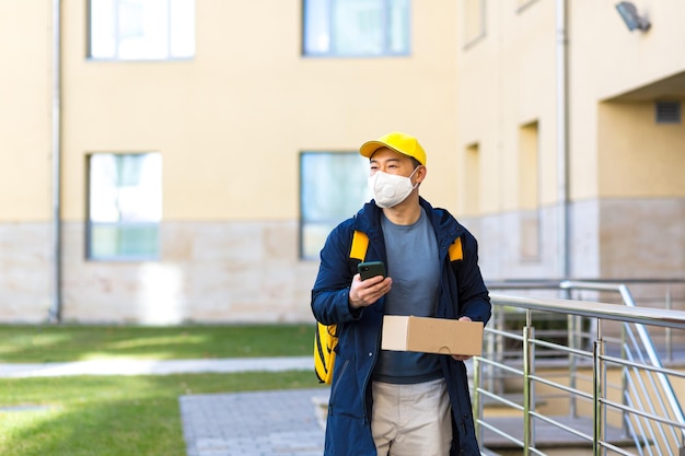 Asian male food courier in Yellow uniform using mobile app on smartphone while walking down city street with insulated with thermal backpack delivering food looking customer address purchase online