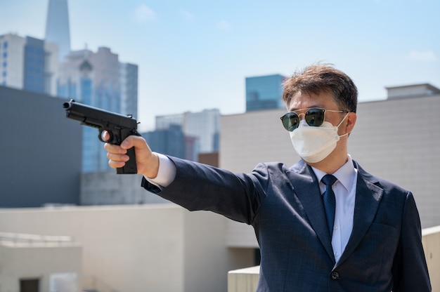 An Asian male holding a pistol wearing sunglasses and a mask in the city.