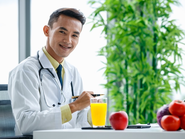 Nutrizionista maschio asiatico che stringe le mani e guarda la telecamera mentre è seduto alla scrivania con cibo sano nell'ufficio dell'ospedale moderno