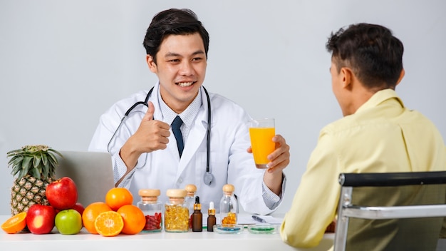 Asian male nutritionist smiling and gesturing thumb up while recommending orange juice to man in clinic