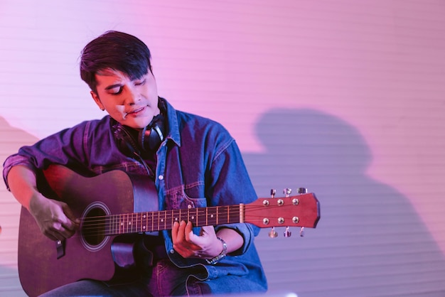 Asian male playing an acoustic guitar places headphones on his neck for listening to the sound in a studio with blue and red lights Rehearsal and recording studio