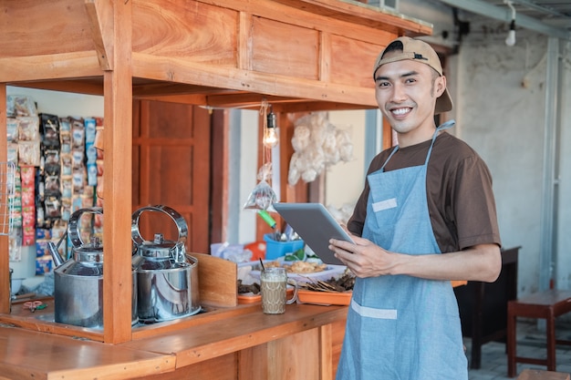 Asian male seller in apron holding a tablet pc standing in side of the cart stall