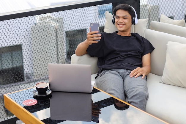 Asian male student relaxing on couch while doing video call using his phone and laptop