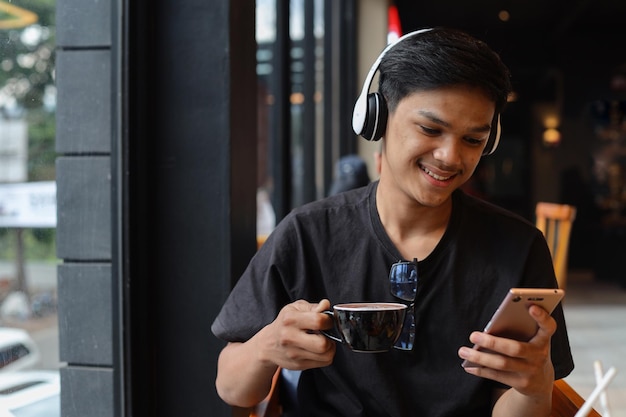 Азиатский студент-мужчина пишет текстовые сообщения на своем телефоне и выпивает чашку кофе в кафе