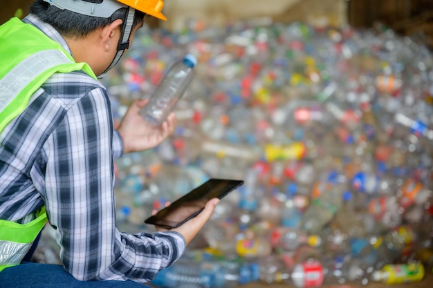 An Asian male worker working at a recycling plant holds plastic bottles and tablets to dispose of and recycle plastic bottles at a small waste recycling plant