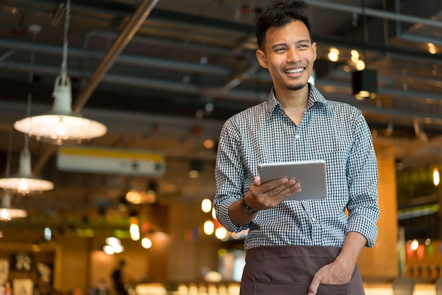 asian man barista holding tablet and looking forward at inside restaurant cafe shop