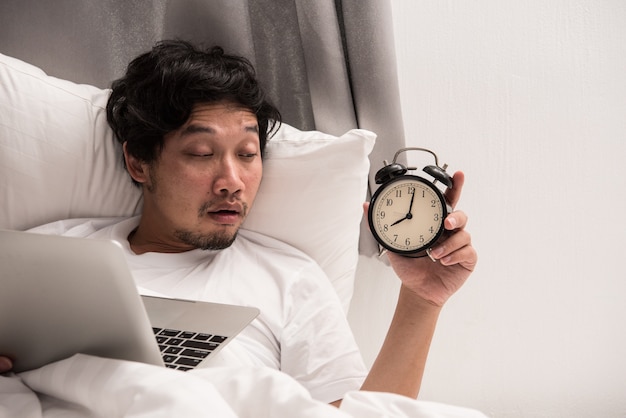 Asian man on bed look on alarm clock when wake up after got sleep while working on computer.