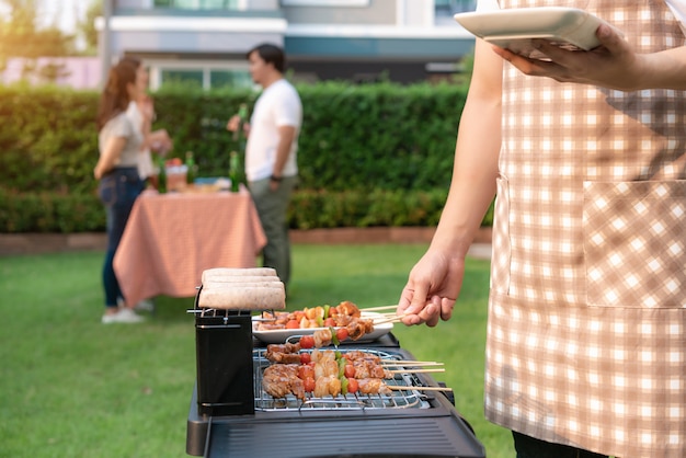 Asian man cooking barbeque grill and sausage for a group of friends to eat party in garden at home.