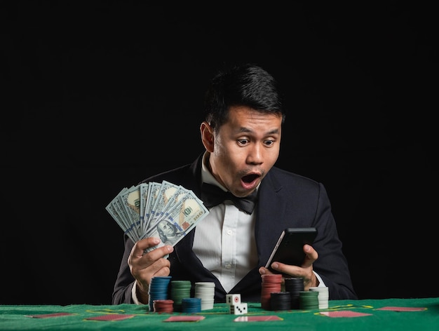 Азиатский дилер или крупье тасует покерные карты, делая ставки в казино, держа в руках мобильный телефон и доллары, счастливая захватывающая улыбка, веселая на черном фоне.