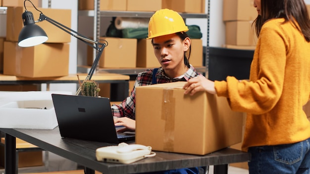 Asian man doing storage room inventory with laptop, organizing merchandise in boxes on shelves. Young adult working in storehouse space to plan order shipment, products distribution.