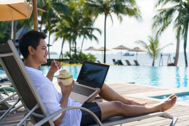 Uomo asiatico che beve acqua di cocco mentre sta lavorando al suo laptop su una piscina con sedie in un resort hotel