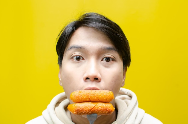 사진 아시아 남자가 노란색 배경에 고립된 포크에 소시지를 먹고 들고 있습니다. 건강에 좋지 않은 패스트푸드 콘크