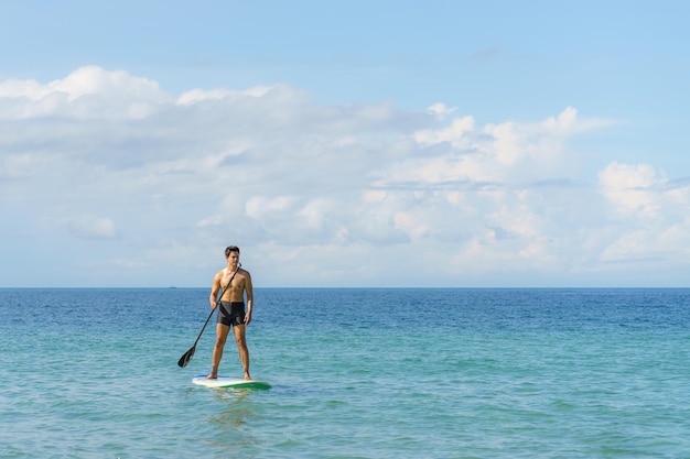 夏の日にターコイズブルーの熱帯の澄んだ海でスタンドアップパドルボードを行使するアジア人男性。