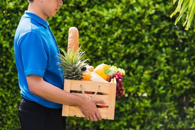 Asian man farmer wears delivery uniform he holding full fresh vegetables and fruits in crate wood box in hands ready give to customer harvest organic food on the garden place green leaves background