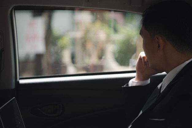 An asian man in a formal suit is in the car he sits behind the\
driver\'s seat and is on his way to work at a company traveling by\
car safe driving respecting traffic rules