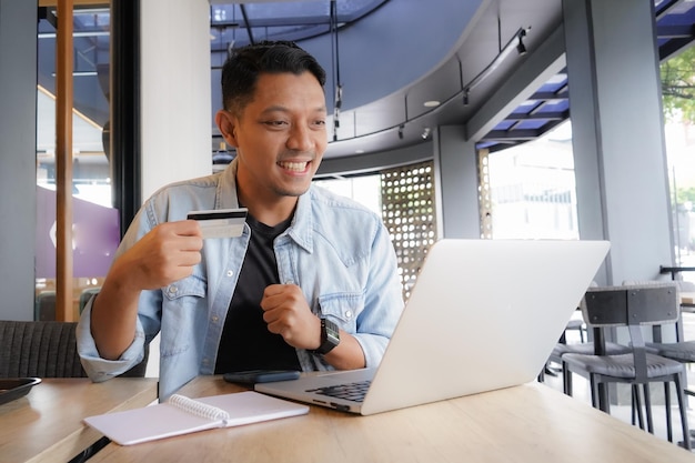 Foto uomo asiatico felice ed entusiasta con camicia blu che usa laptop e cellulare in un caffè online