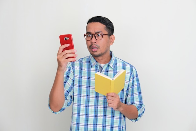 Asian man holding a book showing shocked expression when looking to his handphone