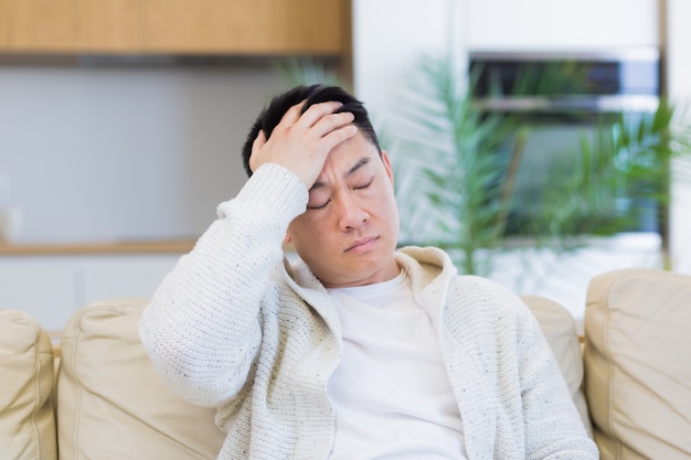 asian man holding his head with a severe headache at home in a room on the couch
