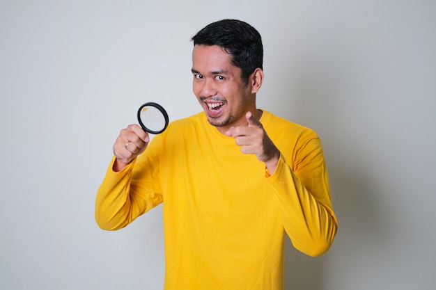 Asian man holding magnifying glass and showing funny expression while pointing forward