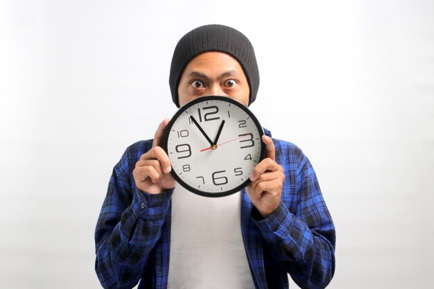 an Asian man holds a clock in front of his face looking at the camera with a surprised expression