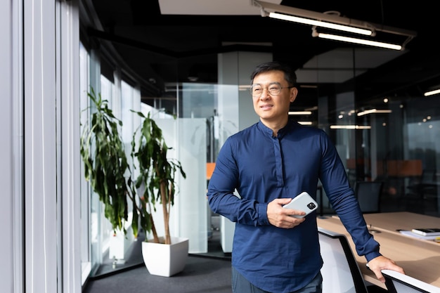 Asian man inside office standing near window dreaming and smiling businessman in shirt and glasses