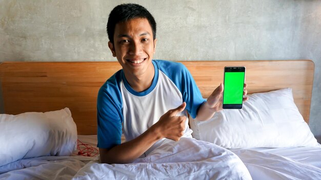 L'uomo asiatico sta tenendo un handphone dello schermo verde nella camera da letto