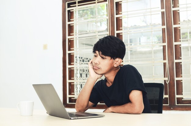 An Asian man is sitting in front of the laptop with a thoughtful expression