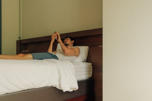 Asian man is using smartphone on the bed in the hotel. Concept of laziness and relax.