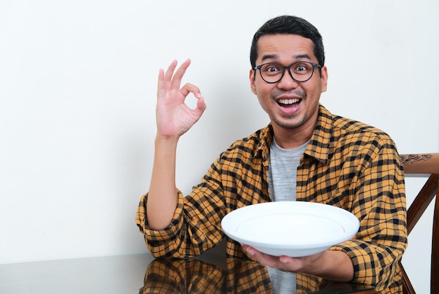 Asian man looking camera with excited expression showing empty dinner plate