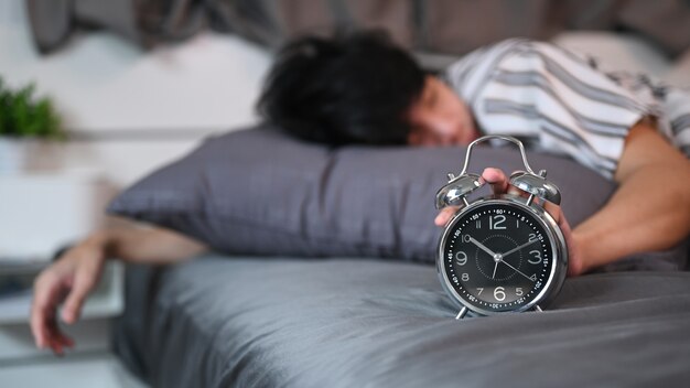asian man lying on his bed and stopping alarm clock.