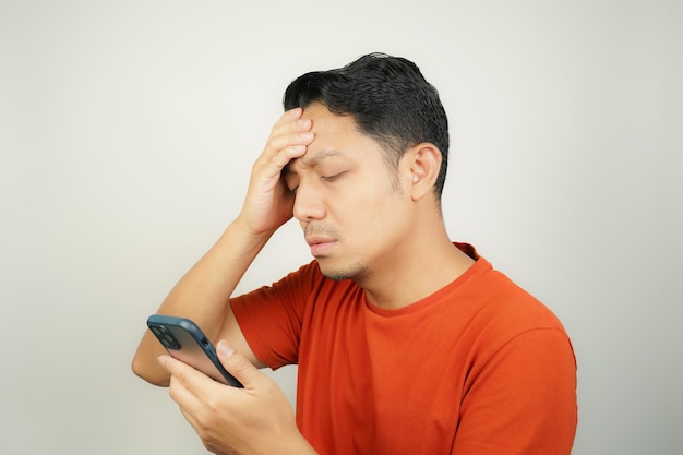 Photo asian man in orange tshirt seen holding head holding problems looking at message on smartphone