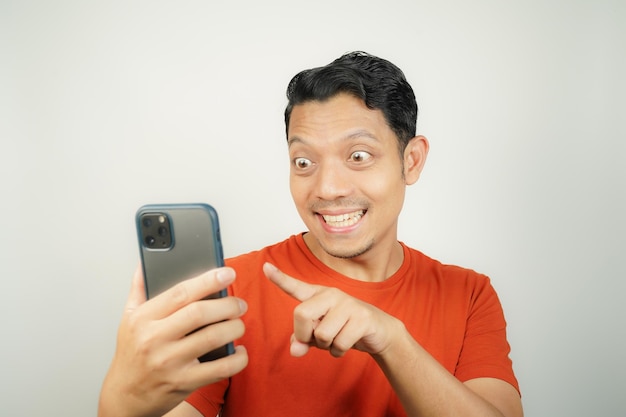 オレンジ色のTシャツを着たアジア人男性が孤立した背景のスマートフォンを見て幸せに笑顔を浮かべています