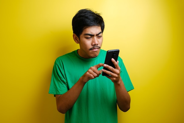 Asian man plays mobile game on his smartphone with serious or angry expression against yellow background