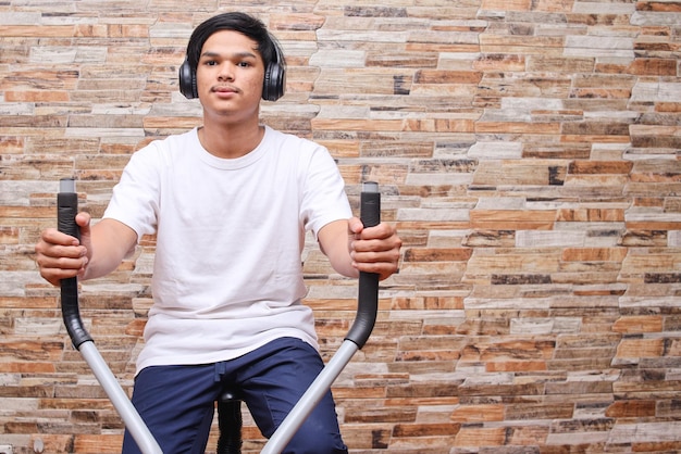 Asian man practicing sport with the exercise bike while using headphones listening to music at home